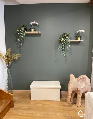 A room with a grey wall, wooden floor, a white bench, and two shelves with plants and an elephant sculpture. - Parifou Air-conditioned cosy house with play area for families (Riom)