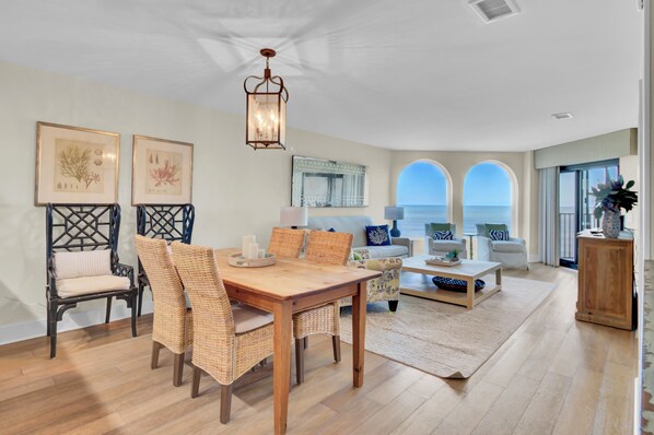 A spacious living room with a wooden dining table, wicker chairs, a large mirror, and a view of the ocean through arched windows.