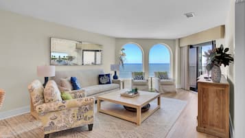 A living room with a sofa, armchairs, a coffee table, and a view of the ocean through arched windows.