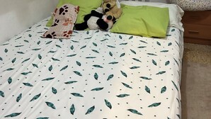 Ein Schlafzimmer mit Bett, Nachttisch, Kommode, einem Fenster mit Jalousien und einem Regal mit diversen Gegenständen.