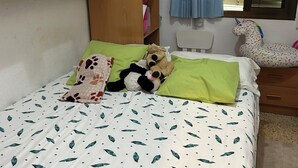 Ein Schlafzimmer mit einem Bett, einem Holzschrank, einem Fenster mit Jalousien und einem Stoffeinhorn-Spielzeug.