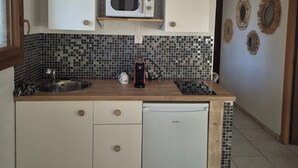 Una cocina compacta con microondas, lavamanos y un refrigerador pequeño. Hay repisas de madera sobre el microondas y una lámpara de techo tejida.