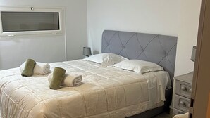 A neatly made bed with a grey headboard, a bedside table with a lamp, and a chair with a blanket. - T4 seafront apartment, magnificent view (Le Lavandou)