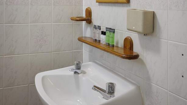 A bathroom with a white sink, wooden shelf, mirror, and wall-mounted soap dispenser. - Hotel Sofia (Blackpool)