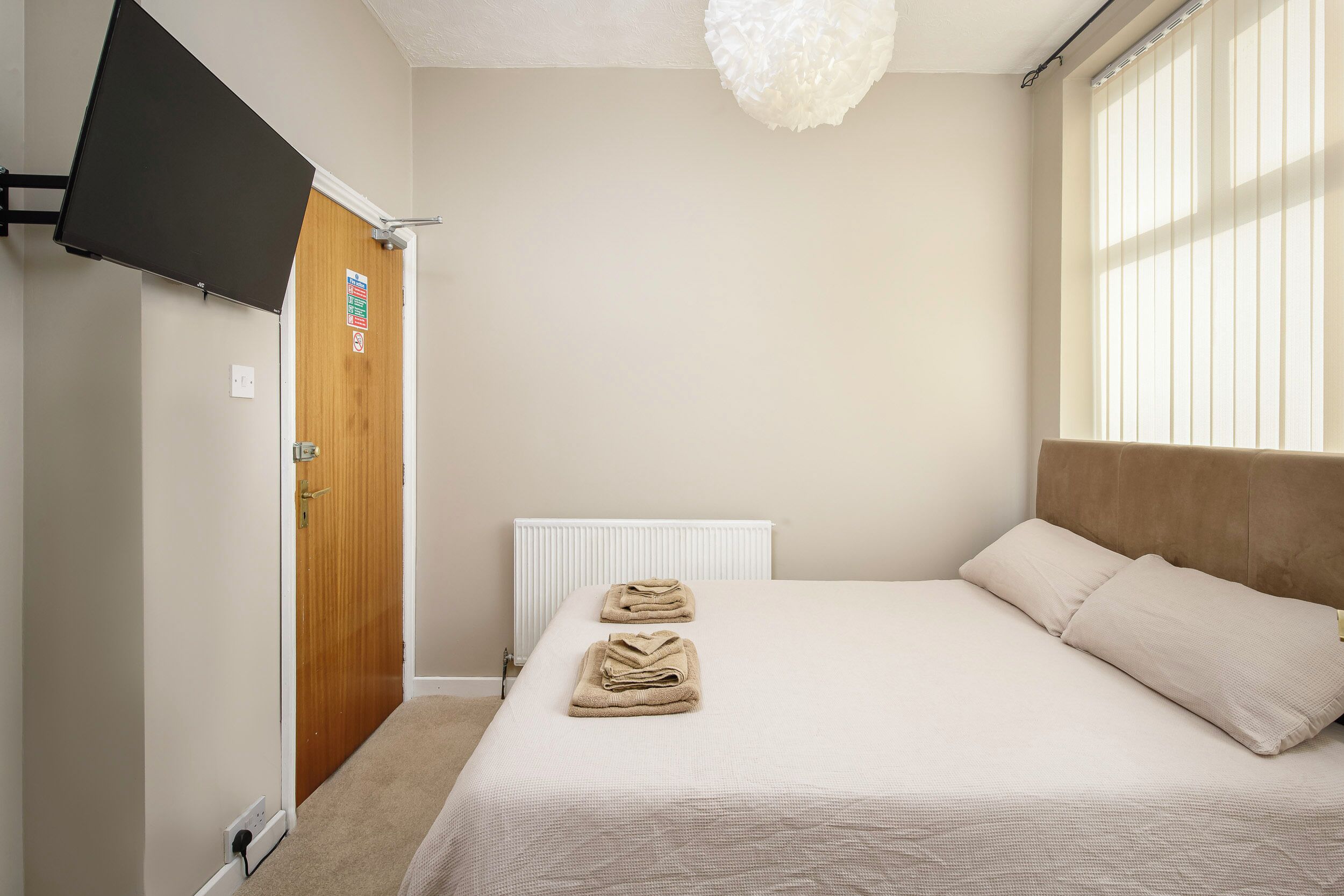 A bedroom with a bed, a wooden door, a white radiator, and a window with blinds.