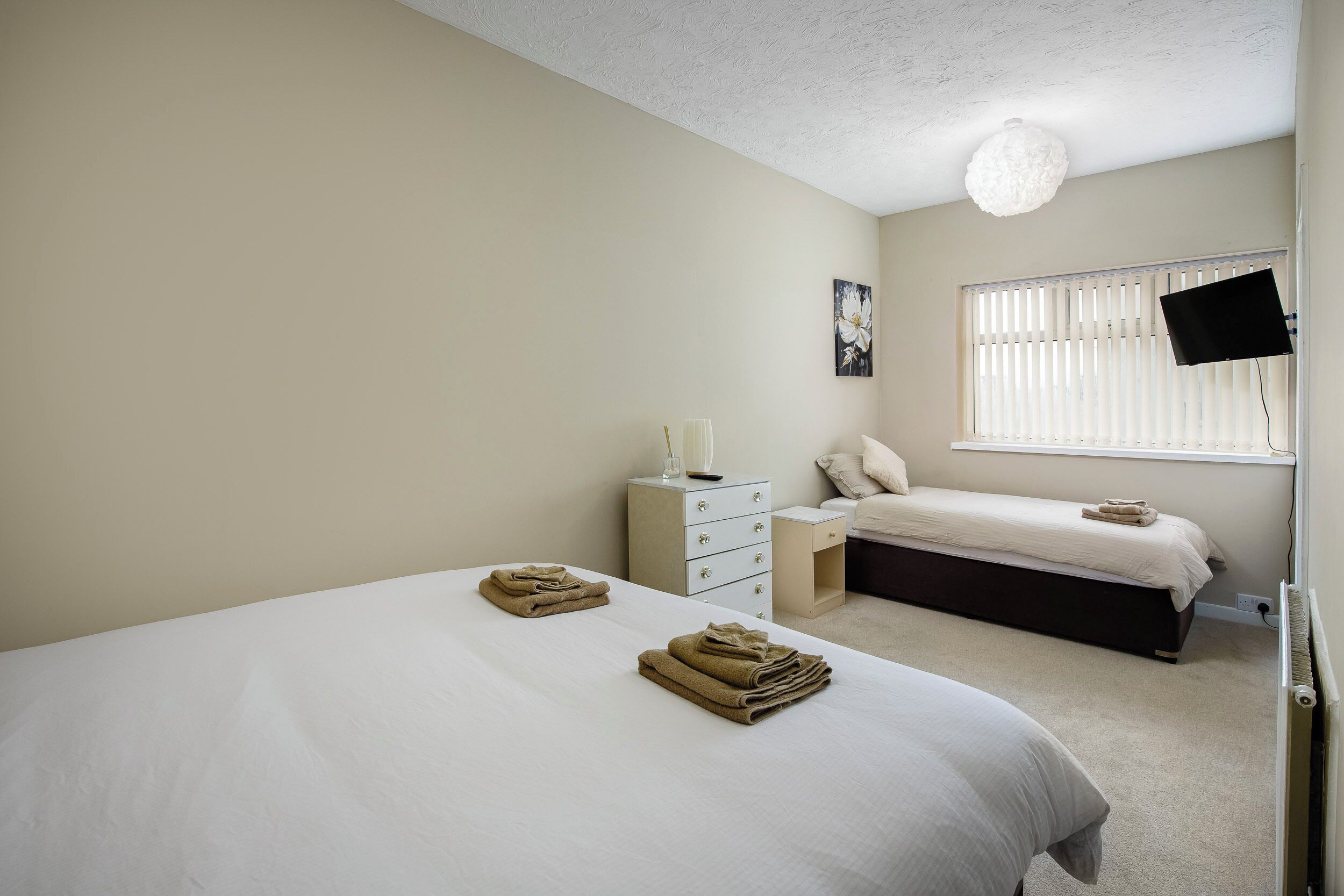 A bedroom with two beds, a bedside table, a nightstand, a TV mounted on the wall, and a window with blinds.