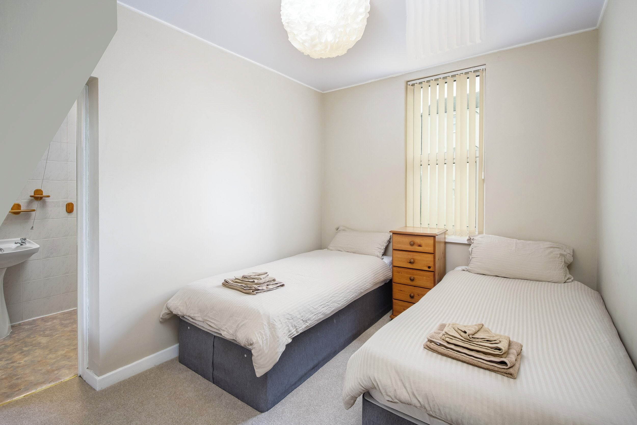 A bedroom with two single beds, a wooden dresser, a window with blinds, and a bathroom with a sink and mirror.