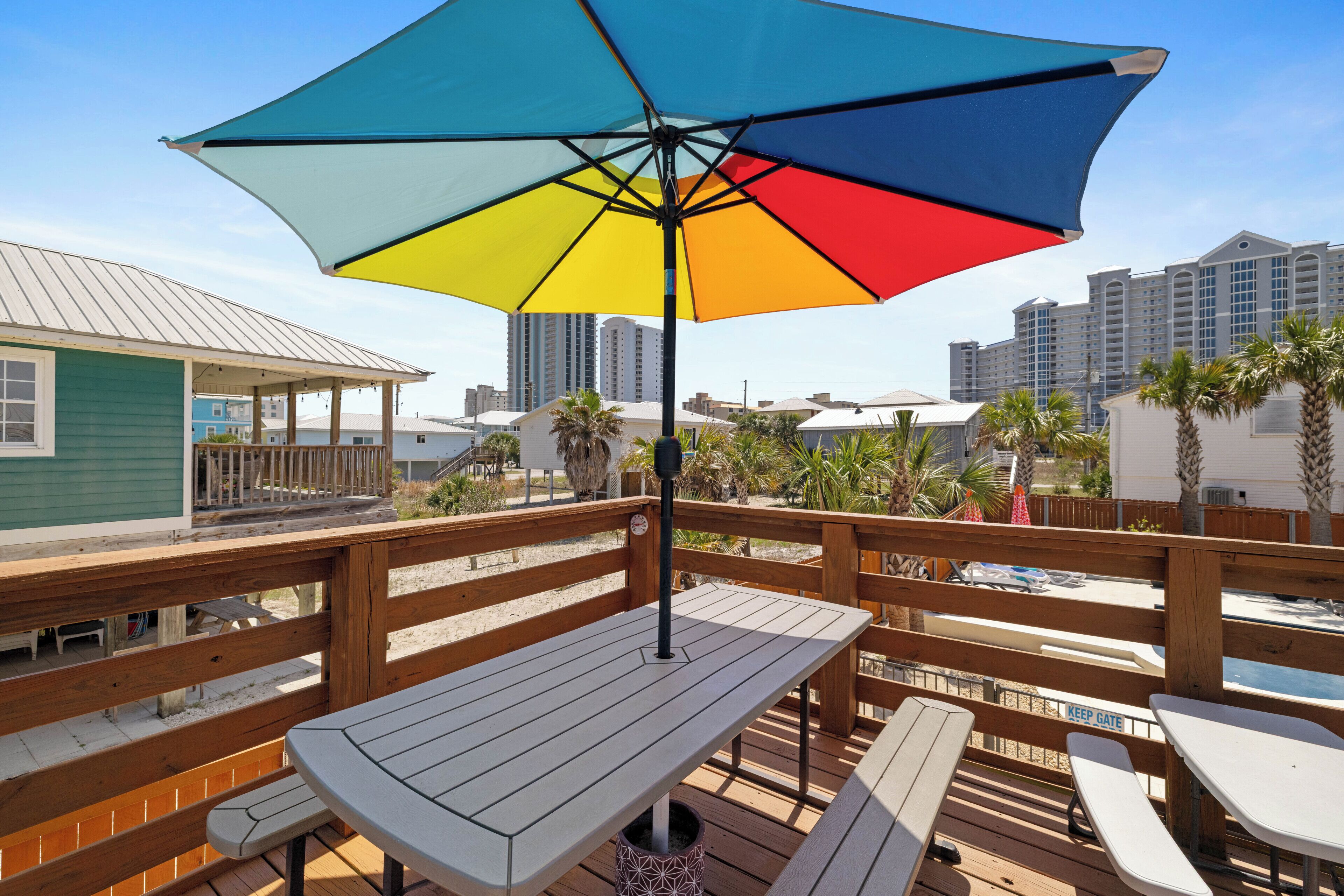 A wooden deck with picnic tables and a colorful umbrella, overlooking a beach with palm trees and a hotel in the background.