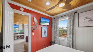 Un dormitorio con cama, televisión, ventana con cortinas, un cuadro y un pasillo que da a una cocina.