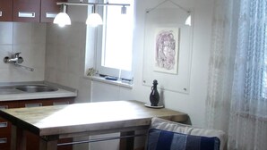 Une cuisine avec un plan de travail en bois, un évier et des placards. Il y a une fenêtre avec un rideau, un vase sur le plan de travail et un coussin sur une chaise.