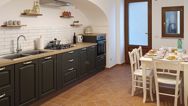A kitchen with dark cabinets, a tiled backsplash, and a dining area with white chairs and a table. - Apartment in the heart of the Amalfi Coast (Praiano)