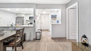 Une cuisine moderne avec des meubles blancs, un coin repas doté d’une table et de chaises en bois, et un couloir menant à une porte.