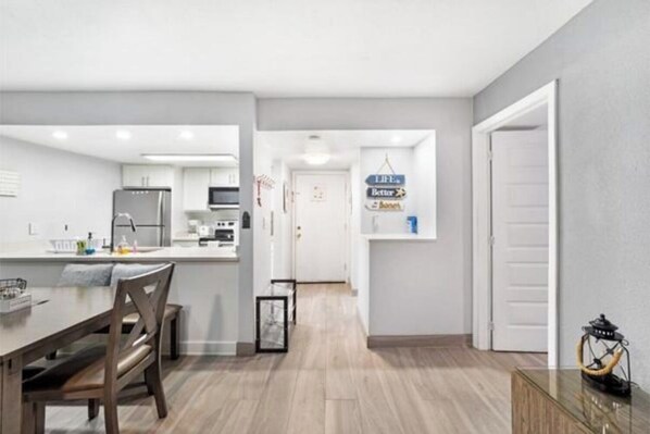 A modern kitchen with white cabinets, a dining area with a wooden table and chairs, and a hallway leading to a door.