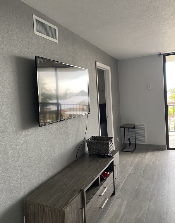 A modern living room with a flat-screen TV mounted on the wall, a wooden console table, and a small table with a basket. - Charming 2-bedroom Beach Access Condo in marvelous Freeport, Lucaya (Freeport)