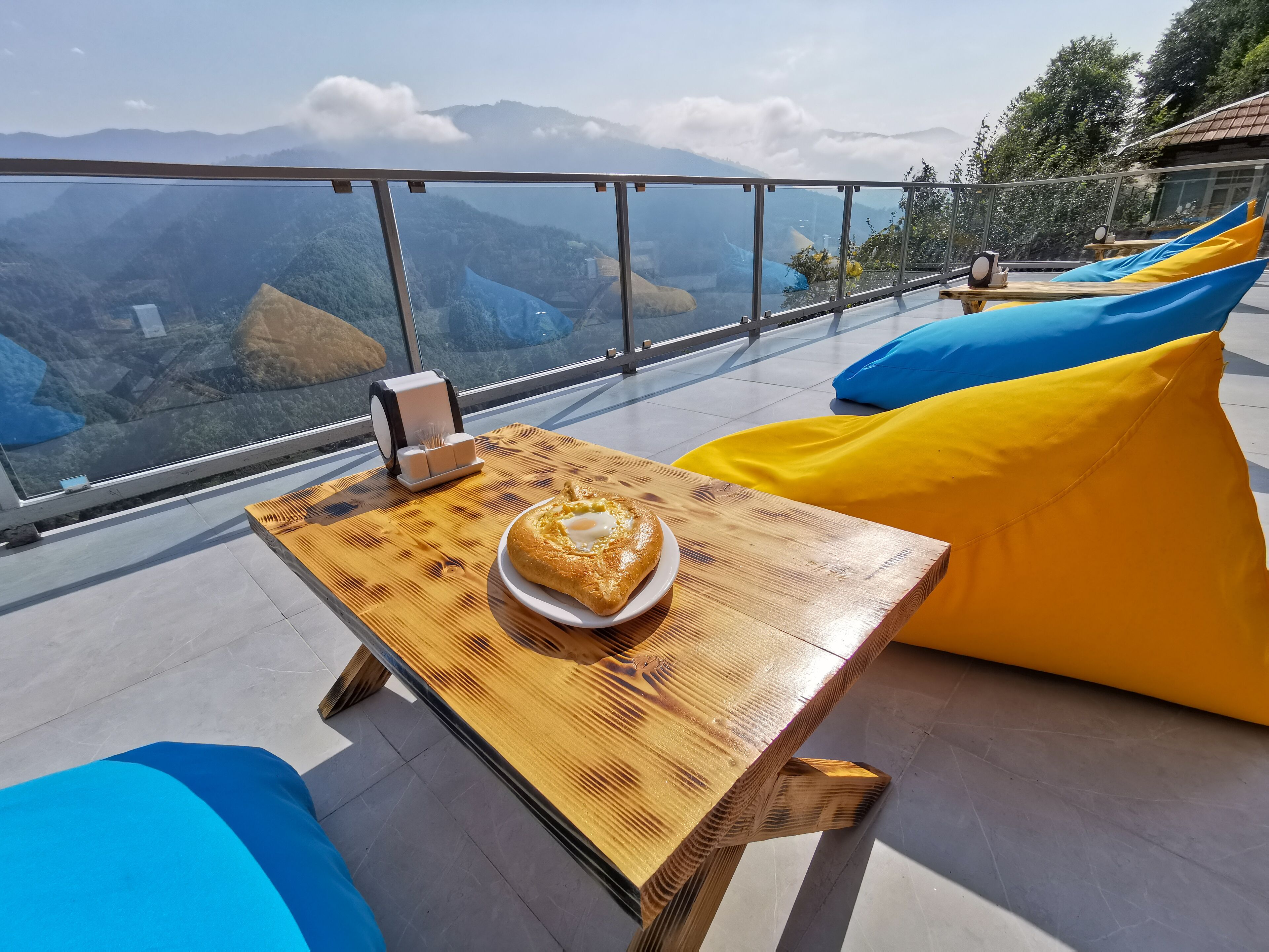 Eine hochgelegene Terrasse mit Holztisch, Speisen auf einem Teller und bunten Sitzsäcken.