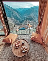 A room with a bed facing a mountainous landscape, a round wooden table with pastries and a cup, and orange cushions.