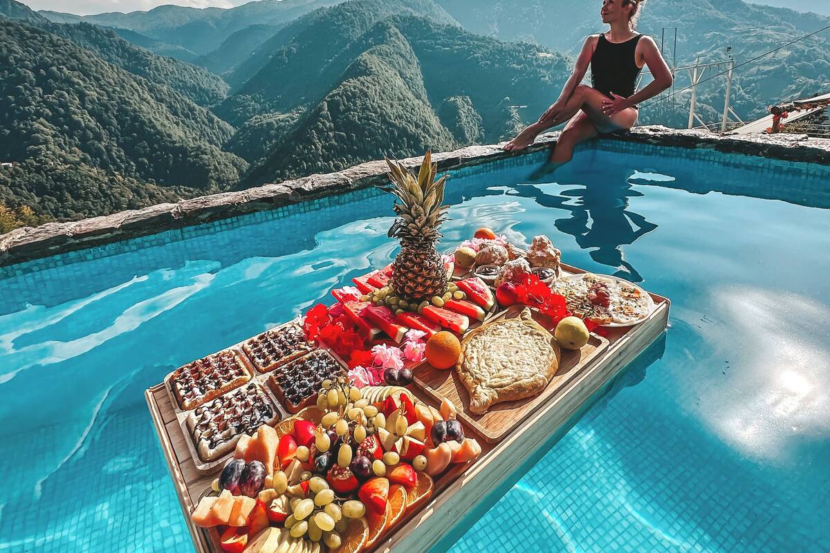 A poolside view with a woman sitting by the pool, surrounded by mountains and a fruit platter.