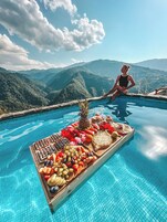 Vista a la piscina con una mujer sentada junto a ella, rodeada de montañas y una bandeja de frutas.