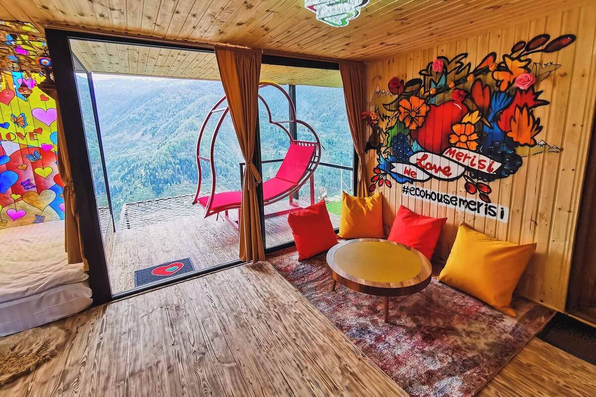 A room with a view of mountains, a swing chair, a round table, and a mural with the text 'Love Merisi we #ecohousemerisi'.