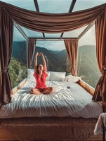 Una cama con vistas a un paisaje montañoso, cortinas y una persona meditando.