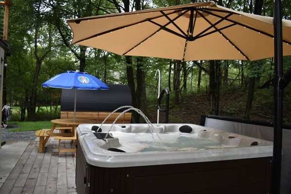 A hot tub with a wooden table and chairs, a blue umbrella, and a barbecue grill in a wooded area. - Aloha Poconos on Sage. (Emerald Lakes)