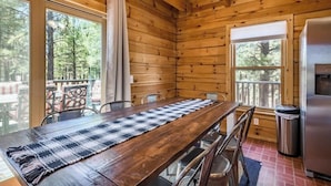A wooden cabin interior with a dining table, chairs, a trash can, and a refrigerator.