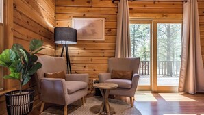 A cozy room with wooden walls, a large window, and a view of trees. There are two armchairs, a small round table, and a floor lamp.