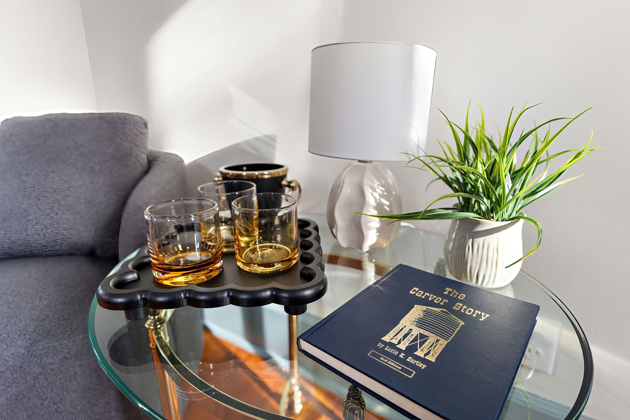 Une table en verre sur laquelle repose un livre intitulé « L’histoire de Carver » et trois verres contenant un liquide ambré.