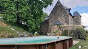 Eine Holzterrasse mit Pool, ein Steinhaus und eine Treppe, die zu einem hügeligen, von Bäumen bewachsenen Hang hinaufführt.