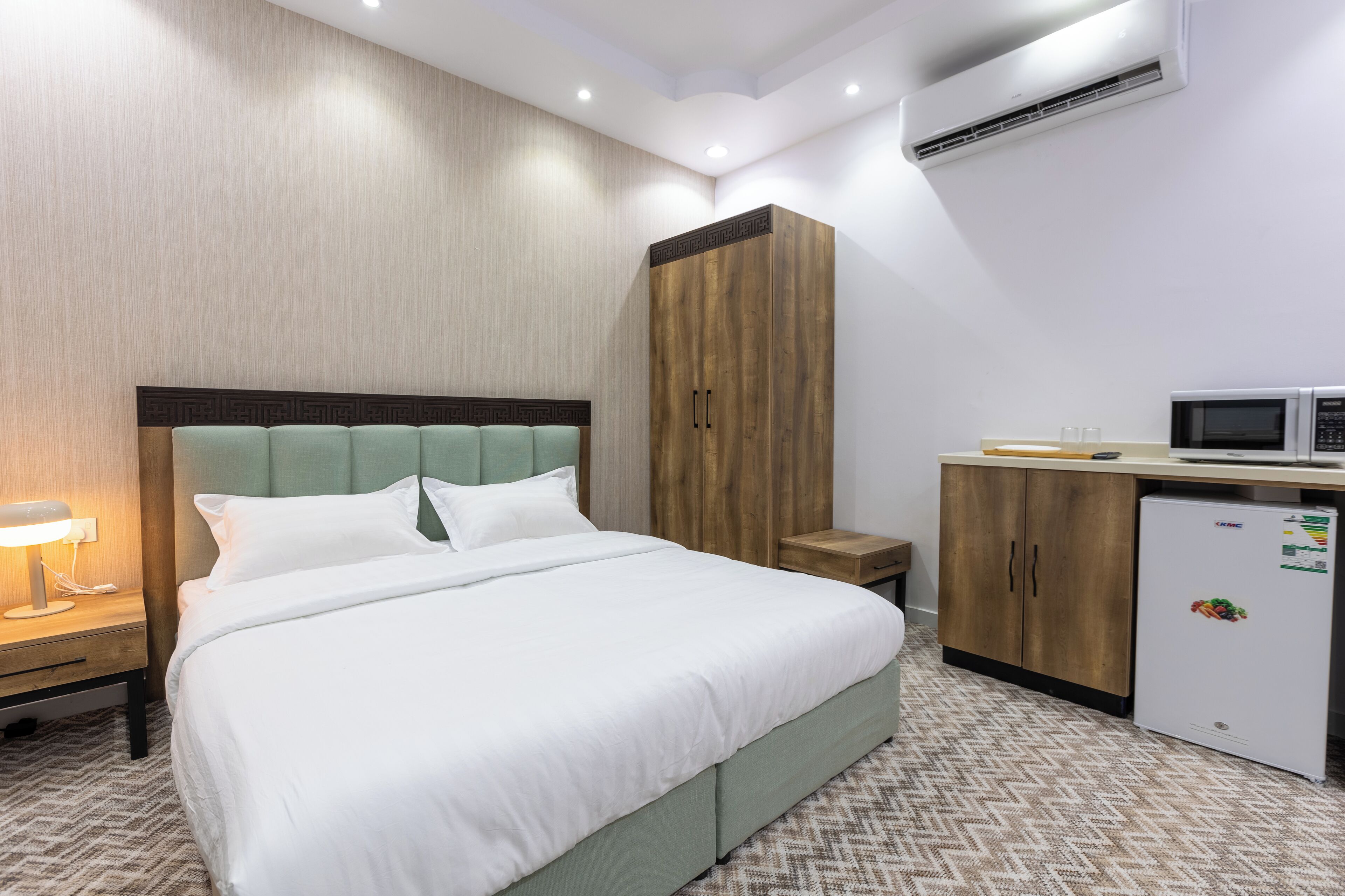 A hotel room with a bed, a wardrobe, a desk with a microwave, and a nightstand with a lamp.