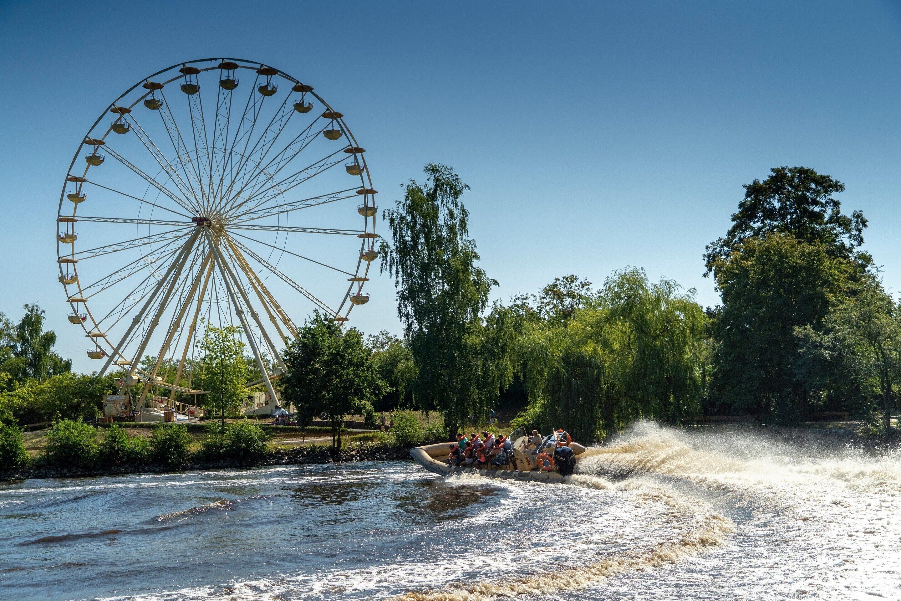 A river with a boat carrying passengers, a large Ferris wheel, and trees in the background.