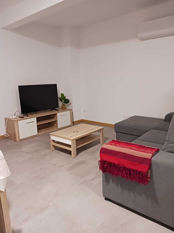 Un salon avec un téléviseur à écran plat posé sur un meuble en bois, un canapé gris, une petite table basse en bois et un plaid rouge.