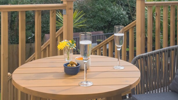 A round wooden table with two glasses of champagne, a small bowl of olives, and a vase with yellow flowers on a deck with wooden railings.