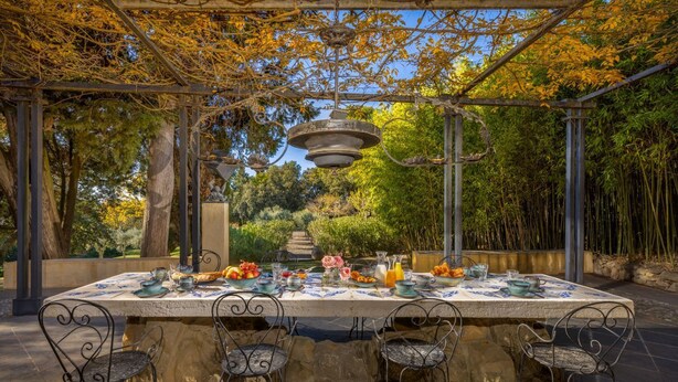 An outdoor dining area with a pergola, stone table, metal chairs, and a garden backdrop.