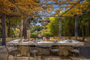 An outdoor dining area with a pergola, stone table, metal chairs, and a garden backdrop.