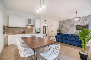Een moderne keuken met witte kasten, een eettafel met witte stoelen, en een blauwe bank in een woonkamer met een groot, bladmotief behang.