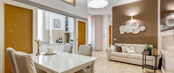 A modern living room with a white sofa, a dining table with chairs, and a kitchen area visible through an open door.