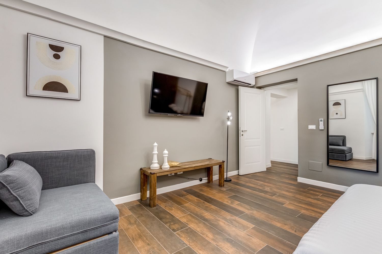 A modern hotel room with a grey sofa, a wooden bench, a flat-screen TV, and a large mirror.