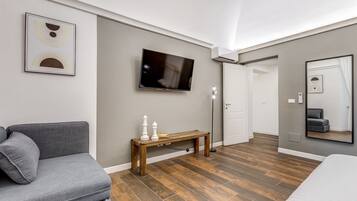 A modern hotel room with a grey sofa, a wooden bench, a flat-screen TV, and a large mirror.