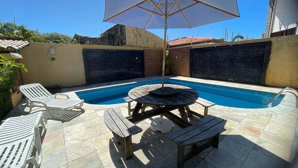 Una piscina con mesa y sillas de madera, rodeada de piso de baldosas y una sombrilla grande.
