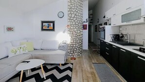 A modern living room with a grey sofa, a round coffee table, and a black and white rug. A stone pillar is in the center, and a kitchen with black cabinets and a white countertop is visible to the right. - Marinero-sea Breeze Escape in Zuljana (Ston)