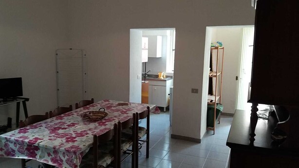 Un comedor con una mesa dispuesta para cuatro, un televisor a la izquierda y una cocina visible a través de un vano abierto.