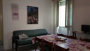 Una habitación con un sofá verde, una mesa de comedor con mantel floreado, un cuadro en la pared y un televisor sobre un mueble.
