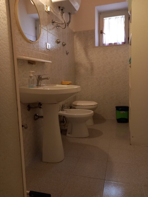 A bathroom with a pedestal sink, two toilets, a mirror, and a window with curtains.