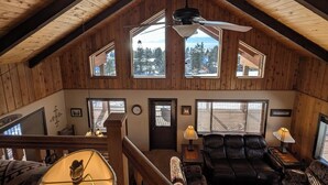 A spacious living room with a high ceiling, wooden walls, and large windows offering a view of the outdoors. - Cedar & Sky Retreat- Rustic Lakeview Cabin (Bigfork)