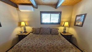 A bedroom with a floral bedspread, two bedside lamps, a window with blinds, and a framed picture on the wall. - Cedar & Sky Retreat- Rustic Lakeview Cabin (Bigfork)