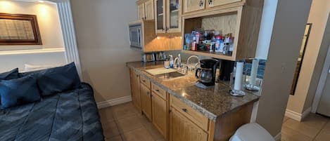 A compact bedroom with a bed, a small kitchenette with a sink, a microwave, and cabinets.