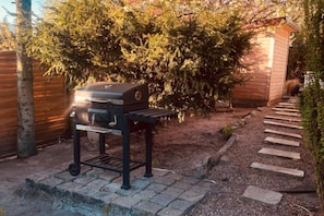 A barbecue grill in a garden with a wooden fence, a wooden shed, and a stone pathway.