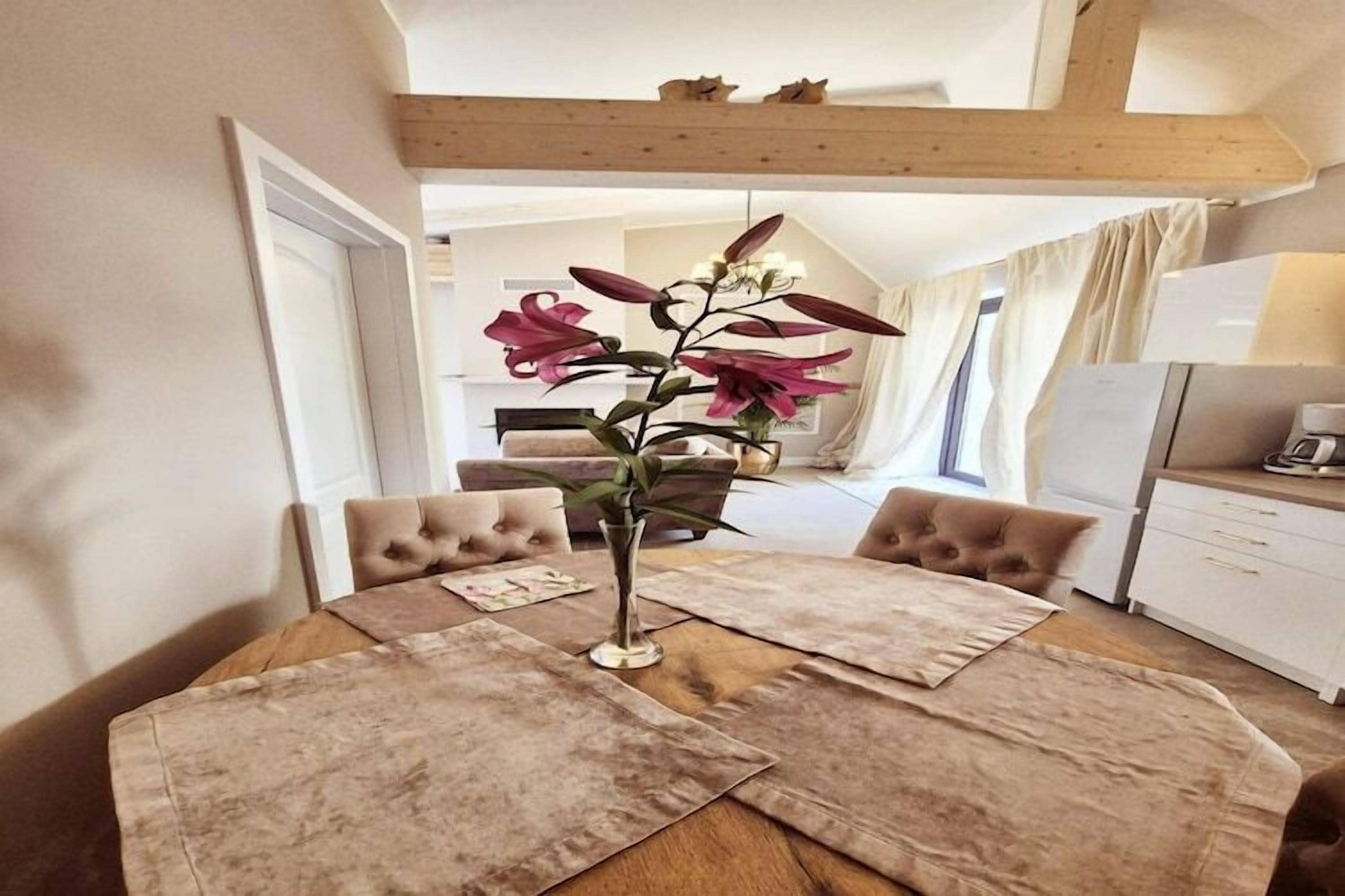 Une table à manger ornée d’un vase de fleurs, entourée de chaises rembourrées, dans une pièce dont le plafond est décoré de poutres en bois, avec un coin cuisine en arrière-plan.