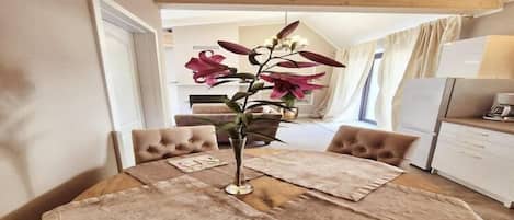 A dining table with a vase of flowers, surrounded by cushioned chairs, in a room with wooden beams on the ceiling and a kitchen area in the background.